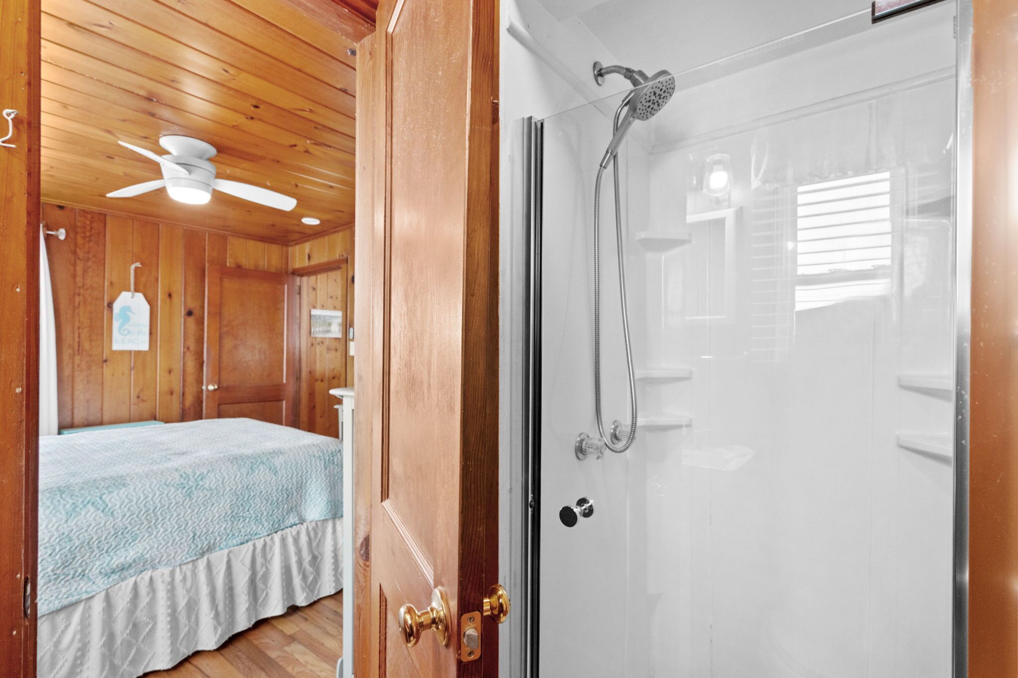 Una habitación de madera con una cama, ventilador de techo y zona de ducha con puerta de cristal.