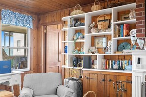 A cozy room with a striped sofa, wooden shelves with books and decorative items, and a window with curtains. - Oceanfront Beach House in Ocean Drive. LOTS of Charm and amazing views! (North Myrtle Beach)