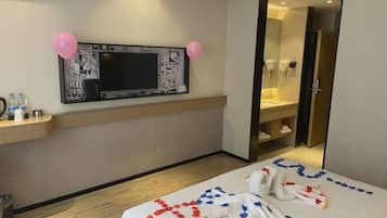 Habitación de hotel con una cama, un televisor en la pared, un baño visible a través de una puerta abierta y un pequeño estante con botellas.