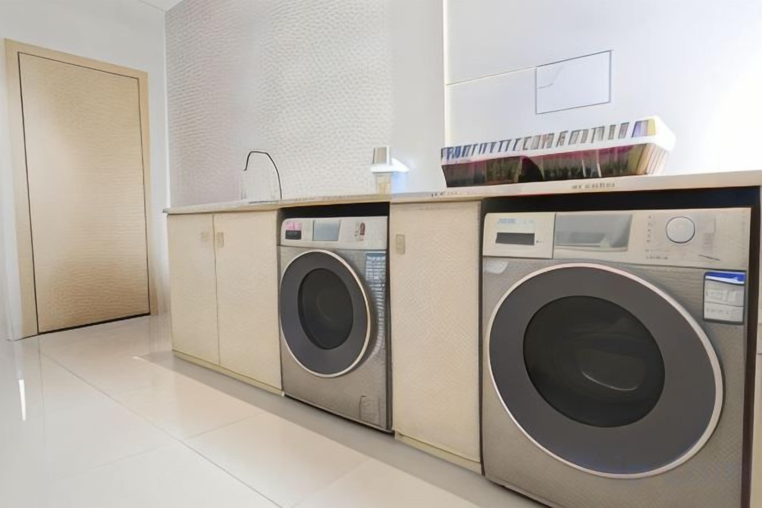 A laundry room with two front-loading washing machines and a dryer, a small table with a lamp, and a door to the left.
