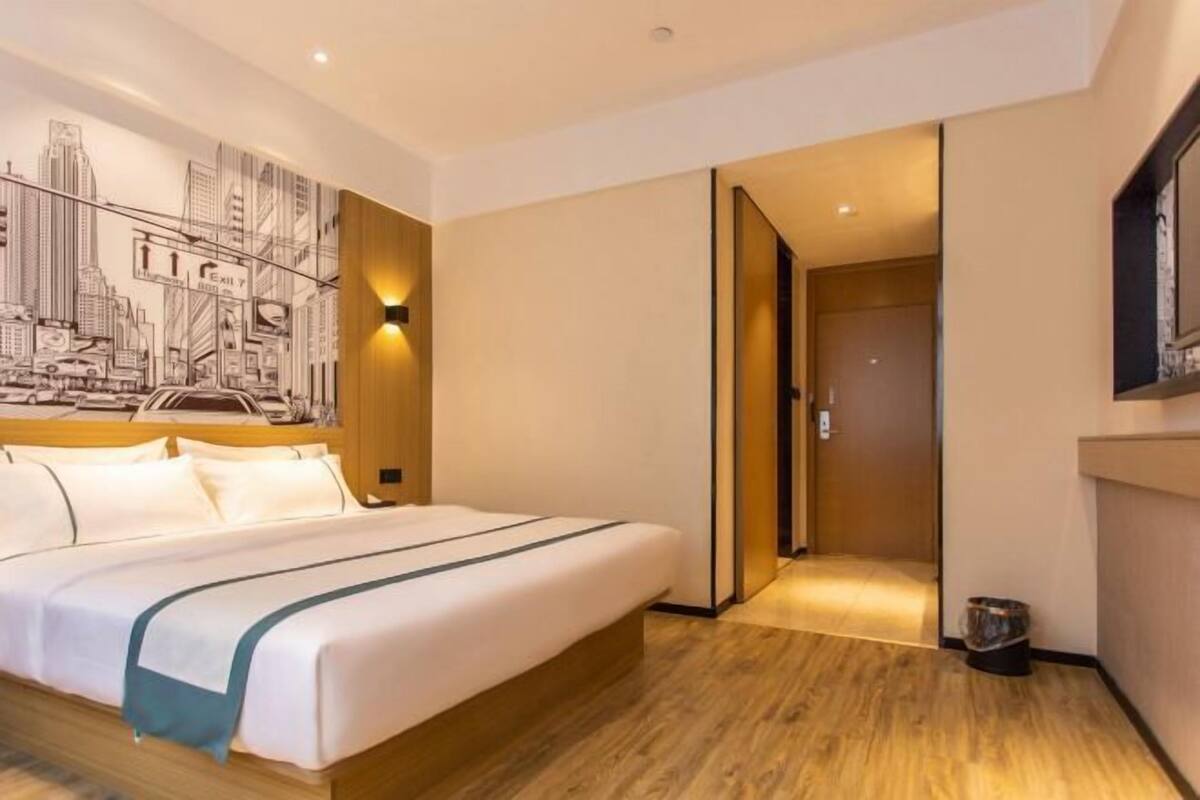 Habitación de hotel con cama, un mural de paisaje urbano, suelo de madera y una puerta que da a otra habitación.