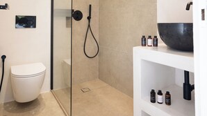 A modern bathroom with a glass-enclosed shower, a white toilet, and a sink area with toiletries. - Loom Suite Ornos Mykonos (Mykonos)
