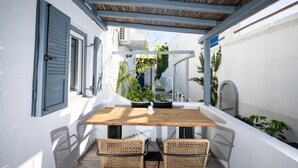A covered outdoor patio with a wooden table and wicker chairs, surrounded by white walls and blue shutters. - Loom Suite Ornos Mykonos (Mykonos)