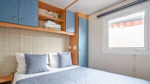 A bedroom with a bed, pillows, a shelf with folded towels, and a window with curtains. - Casa azul (Pataias)