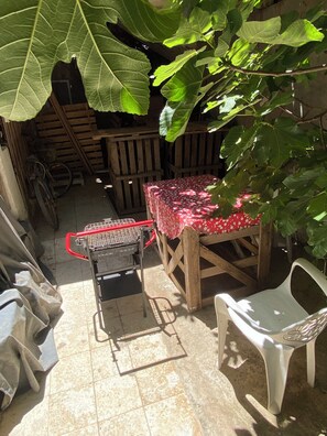A covered outdoor area with a grill, a table with a polka dot tablecloth, a plastic chair, and large green leaves overhead.