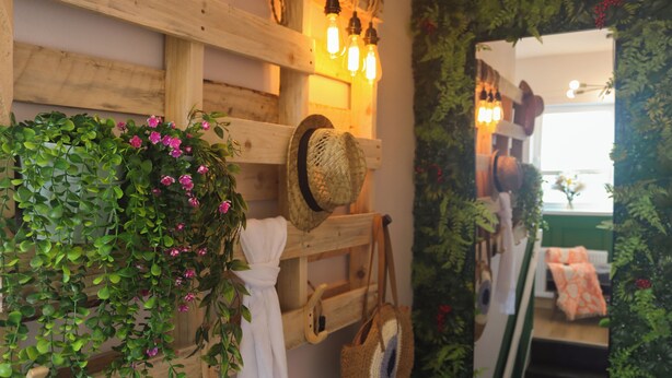 Un intérieur rustique avec un mur en bois, une corbeille suspendue remplie de plantes vertes, une serviette blanche, un chapeau de paille et un sac tressé.