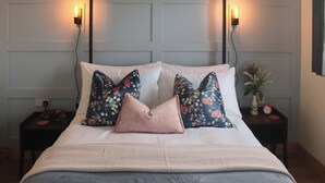 Un lit bien fait, agrémenté d’oreillers à motifs floraux, une table de chevet avec une horloge et des lampes fixées au mur.