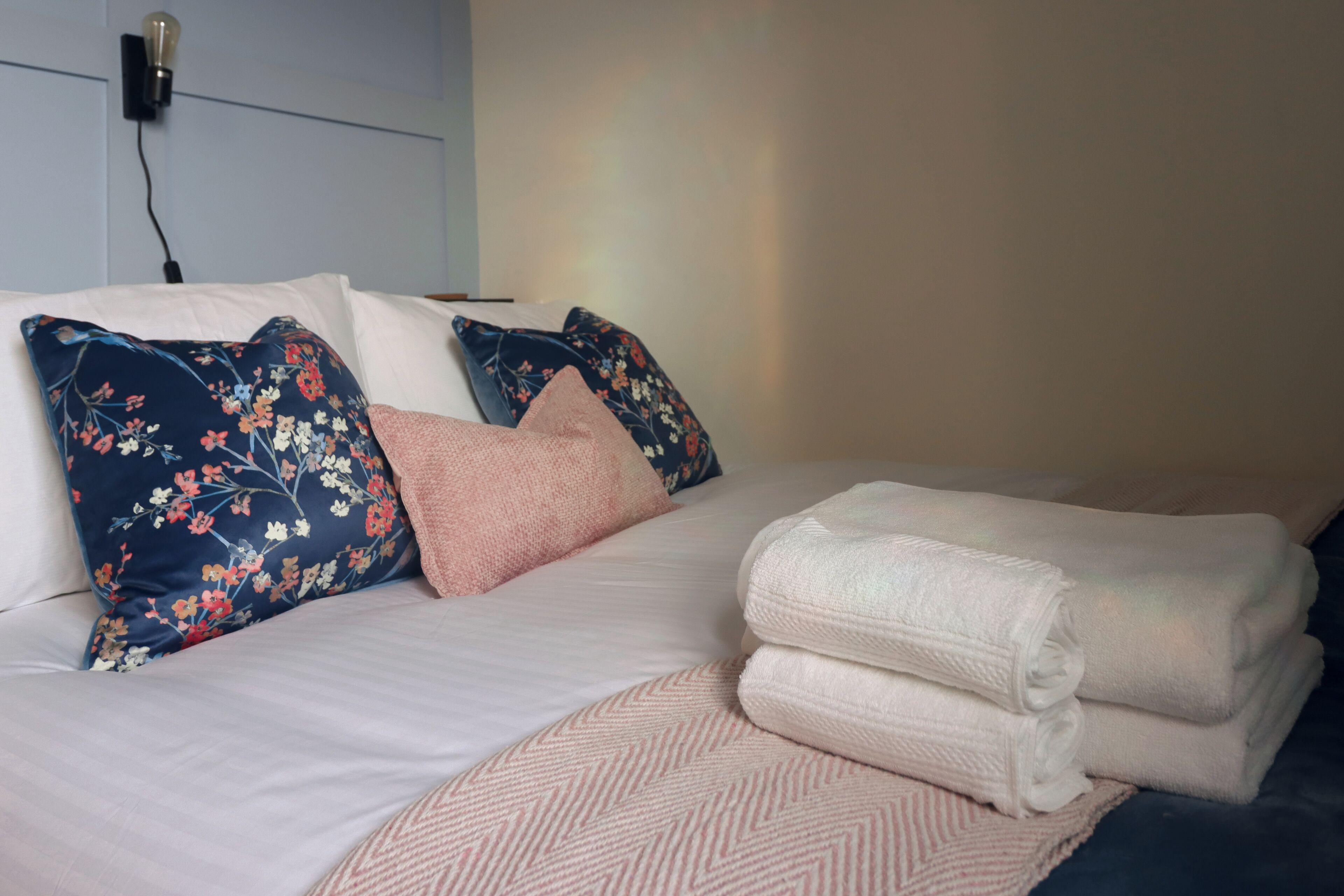 A neatly made bed with floral pillows, a pink throw, and folded white towels.