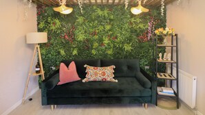 Un salon chaleureux avec un canapé vert foncé, deux coussins décoratifs, une petite table d’appoint avec une lampe et une bibliothèque remplie de livres et d’objets décoratifs.