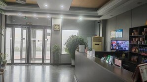 Zona de recepción con puerta de entrada de vidrio, mostrador de recepción, televisor y planta en maceta.