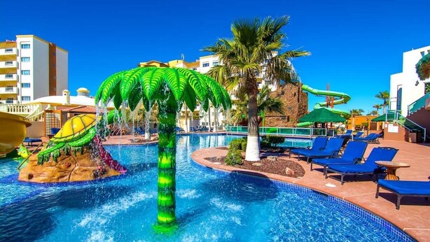 Ein Poolbereich mit einem zentralen Wasserspiel, Liegestühlen und einer Rutsche.