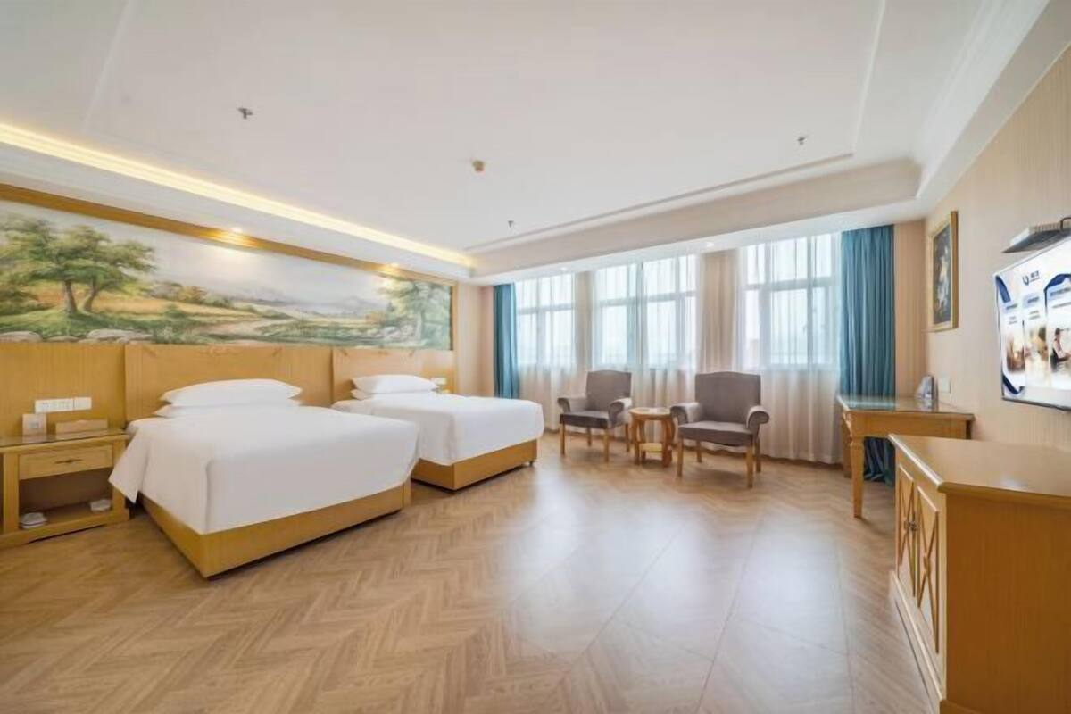 A hotel room with two beds, a desk, a chair, a small table, and a painting on the wall.