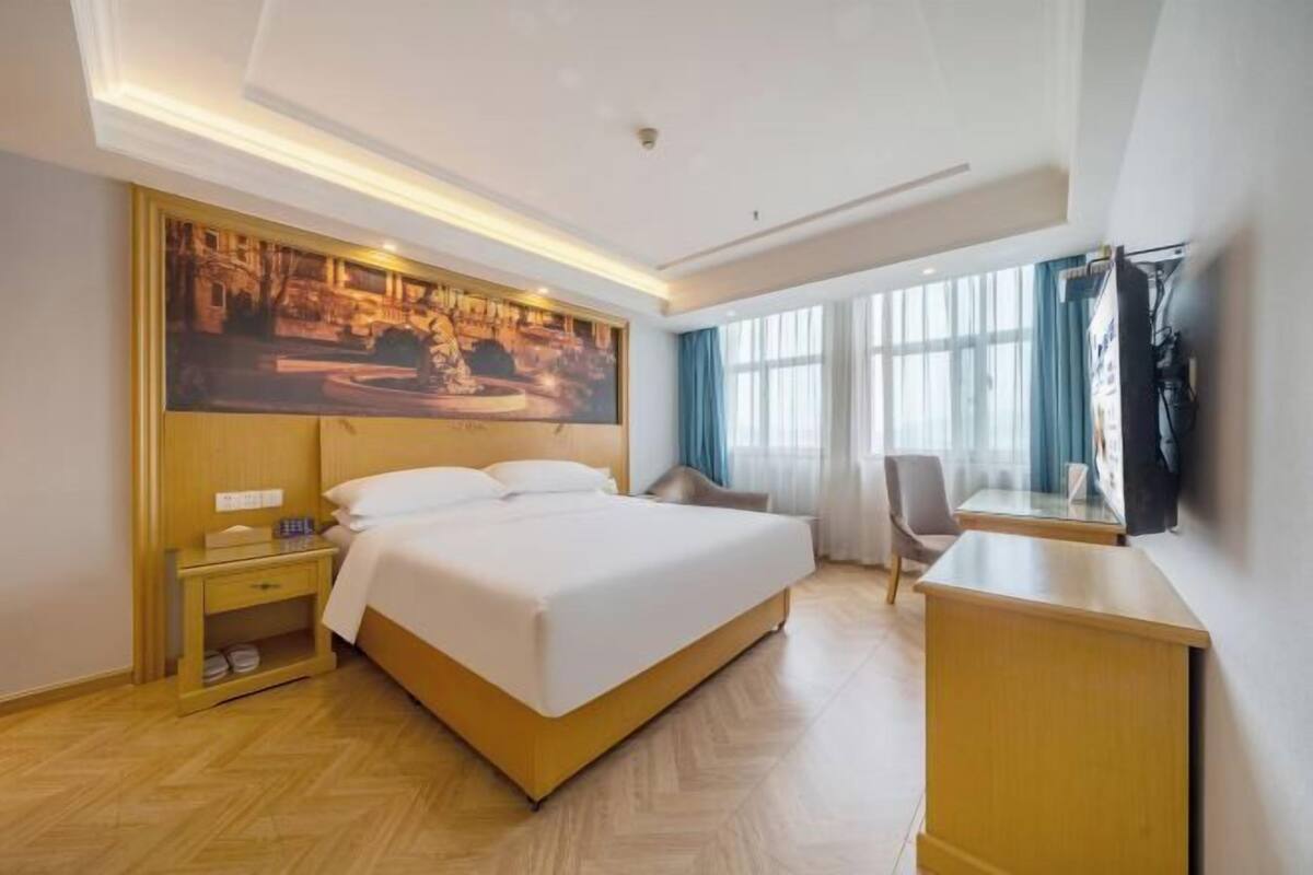 A hotel room with a bed, a desk, a chair, a nightstand, and a large window with curtains.