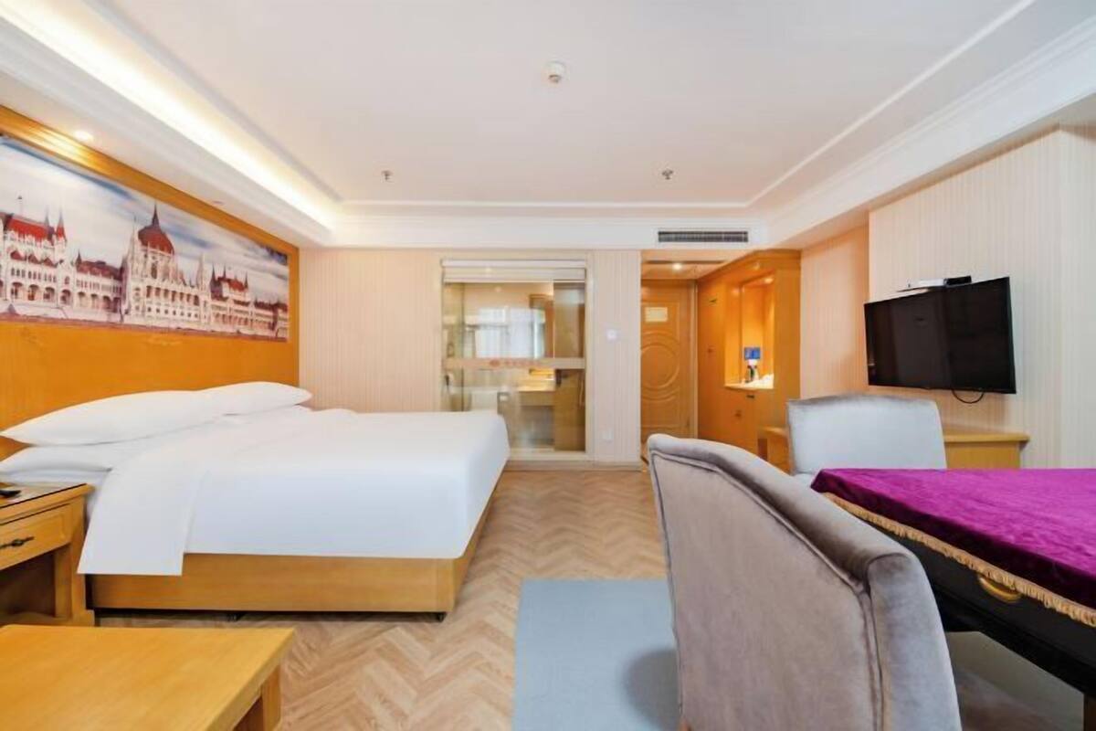 A hotel room with a large bed, a desk, a chair, a TV, and a painting of a building on the wall.