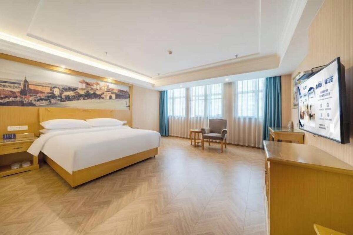 A hotel room with a bed, a chair, a desk, a TV, and a painting on the wall.
