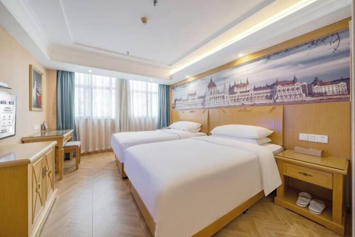 A hotel room with two beds, a desk, and a view of a cityscape.