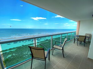 A balcony with two chairs and a table, overlooking a beach with clear turquoise waters and a distant view of the ocean.