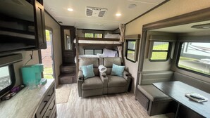 A cozy RV interior with a kitchenette, a seating area with cushions, a TV, and a window with a view of the outdoors.