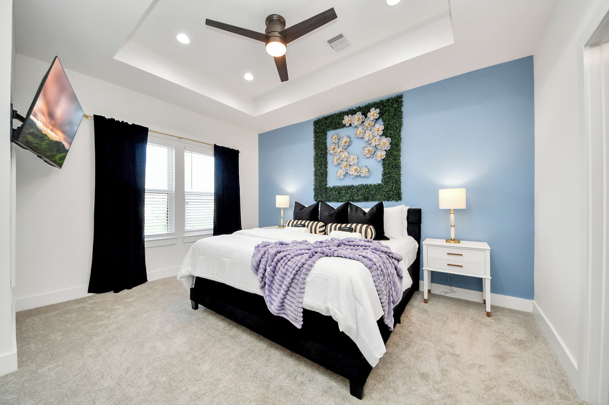 A bedroom with a bed, a ceiling fan, a wall-mounted TV, two bedside tables with lamps, and a decorative wall piece.