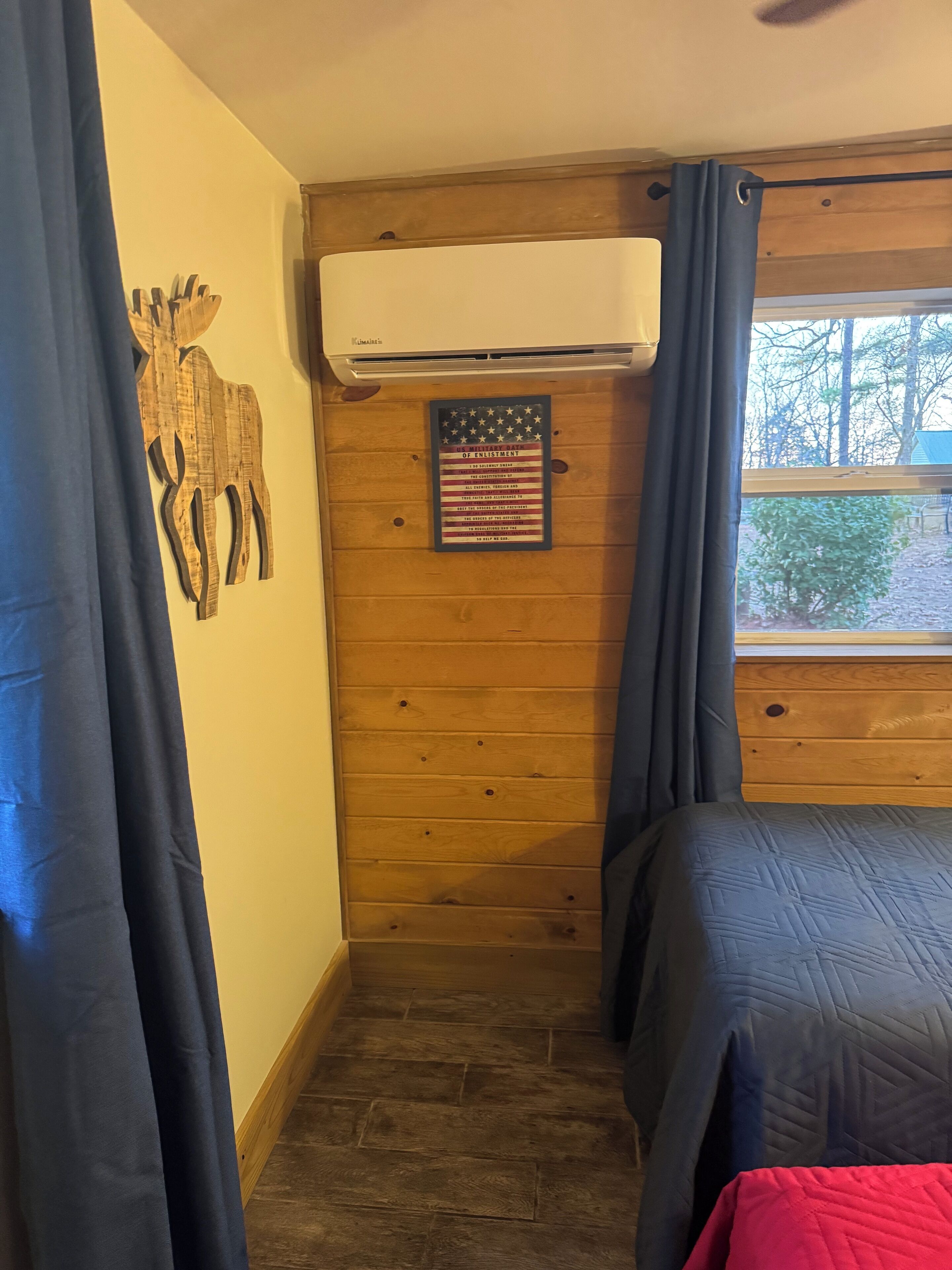 A bedroom with a wooden wall, a mounted air conditioner, a framed American flag, and a window with curtains.
