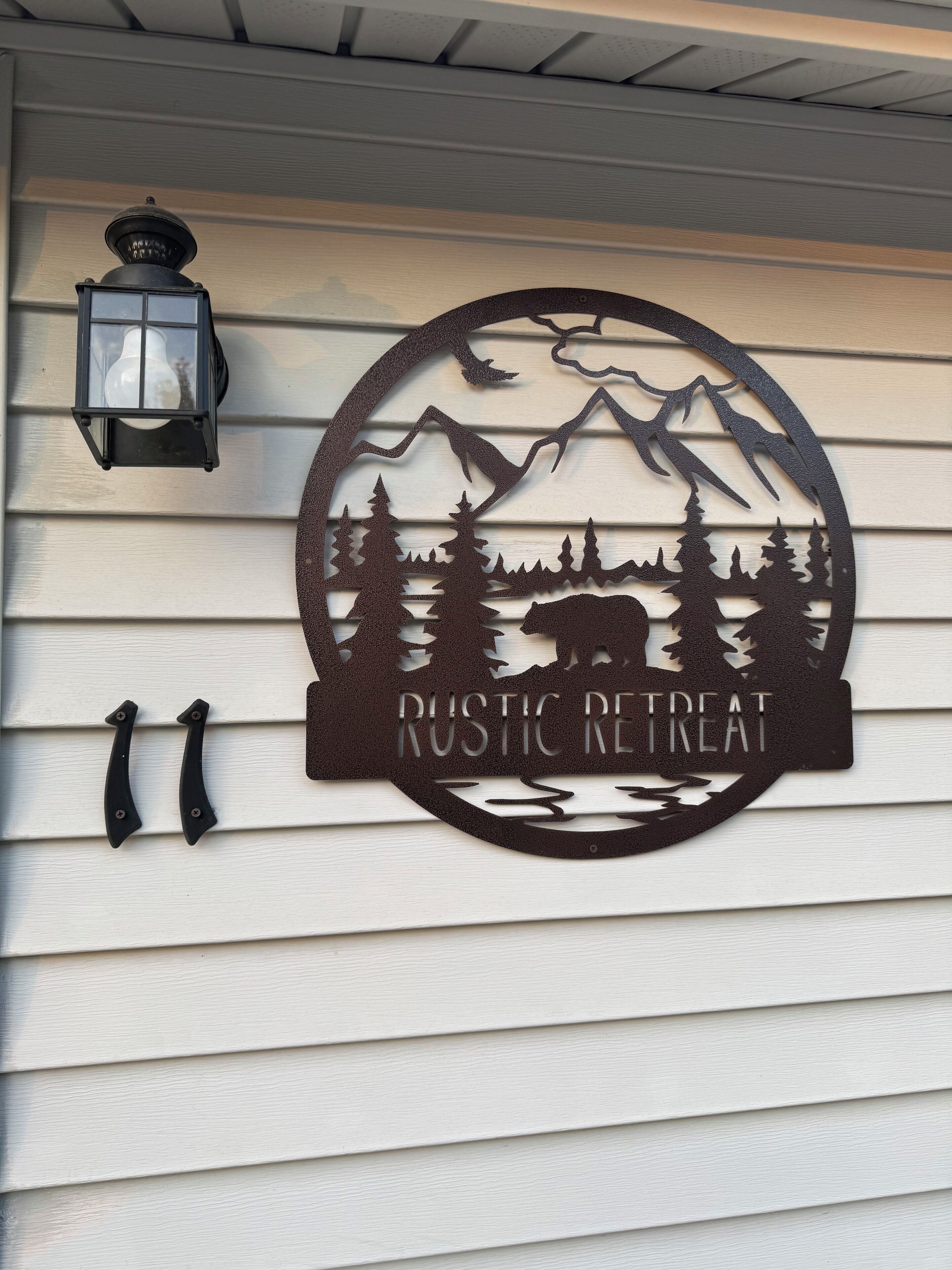 Une enseigne rustique indiquant un lieu de retraite, avec la silhouette d'une montagne et d'une forêt, une lanterne et deux poignées noires sur un mur recouvert de bardage blanc.