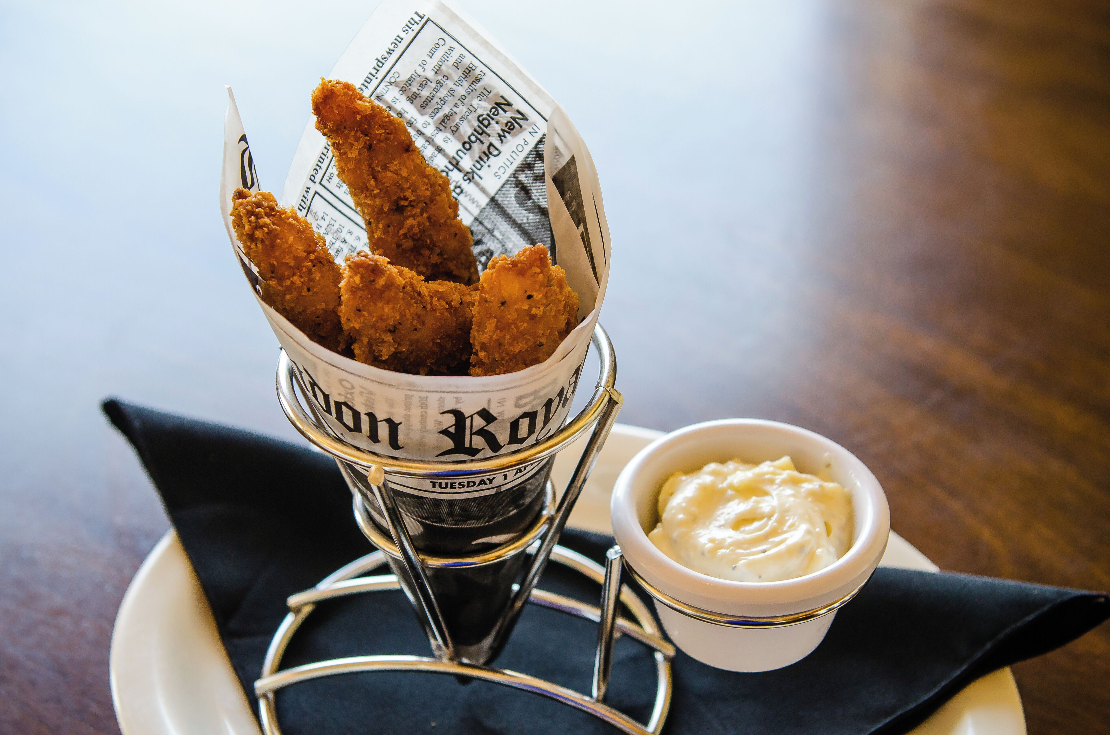 Un cesto de tiras de pollo frito, un periódico y un recipiente pequeño con salsa para mojar, sobre una mesa.