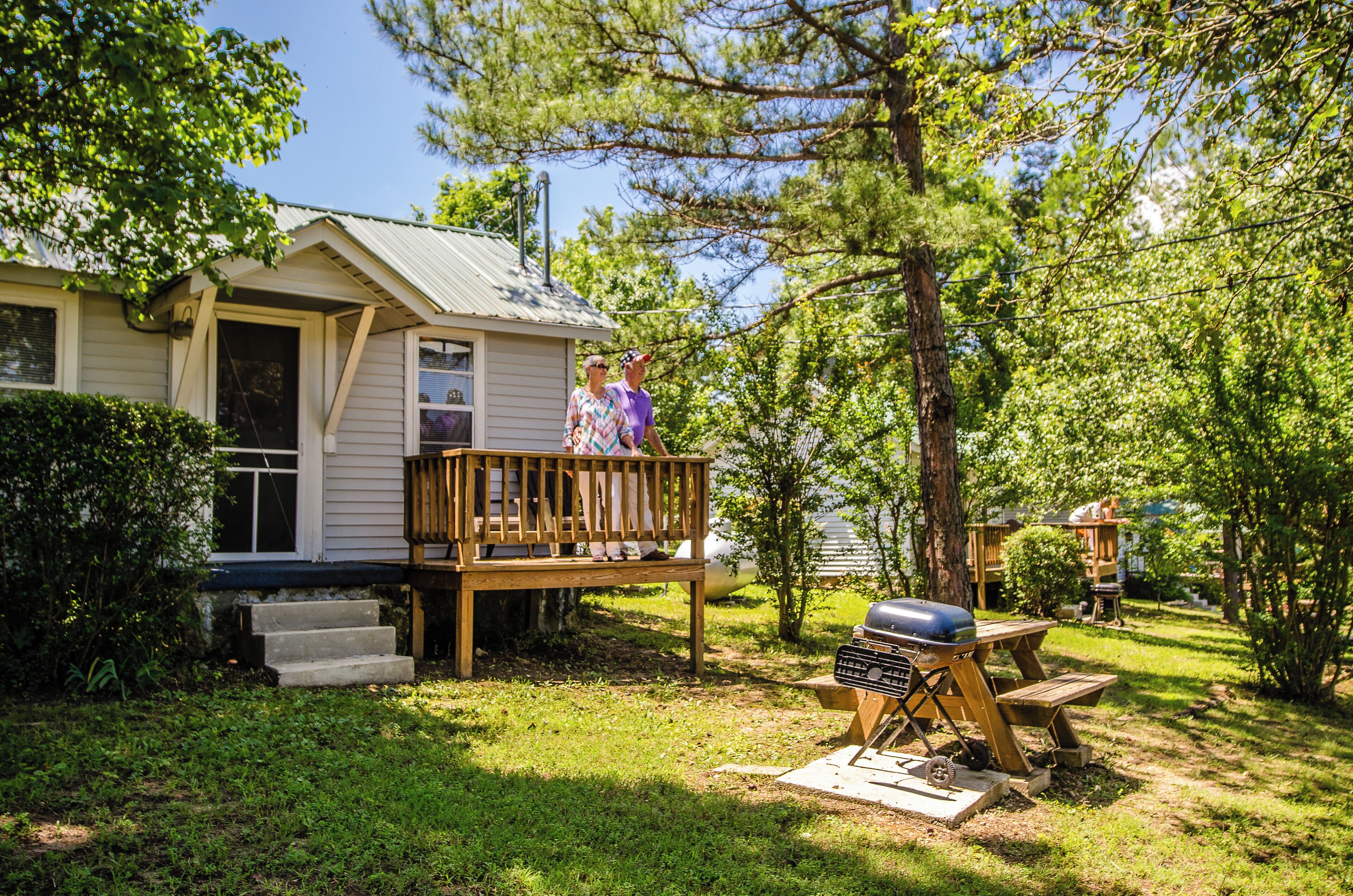 A wooden deck with a barbecue grill, a small house with a porch, and a tree in the background.