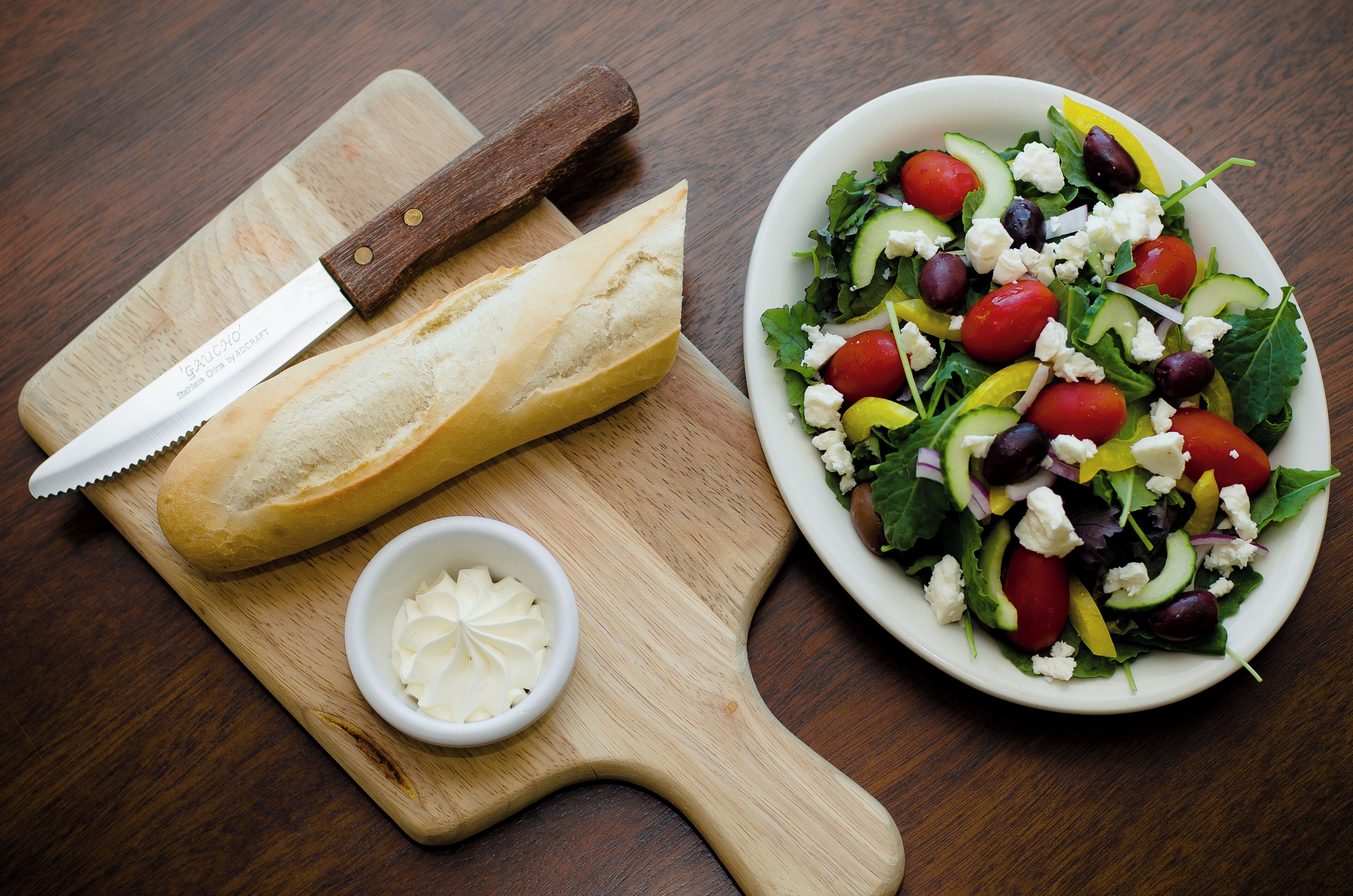 A wooden cutting board with a loaf of bread, a knife, and a small bowl of butter.