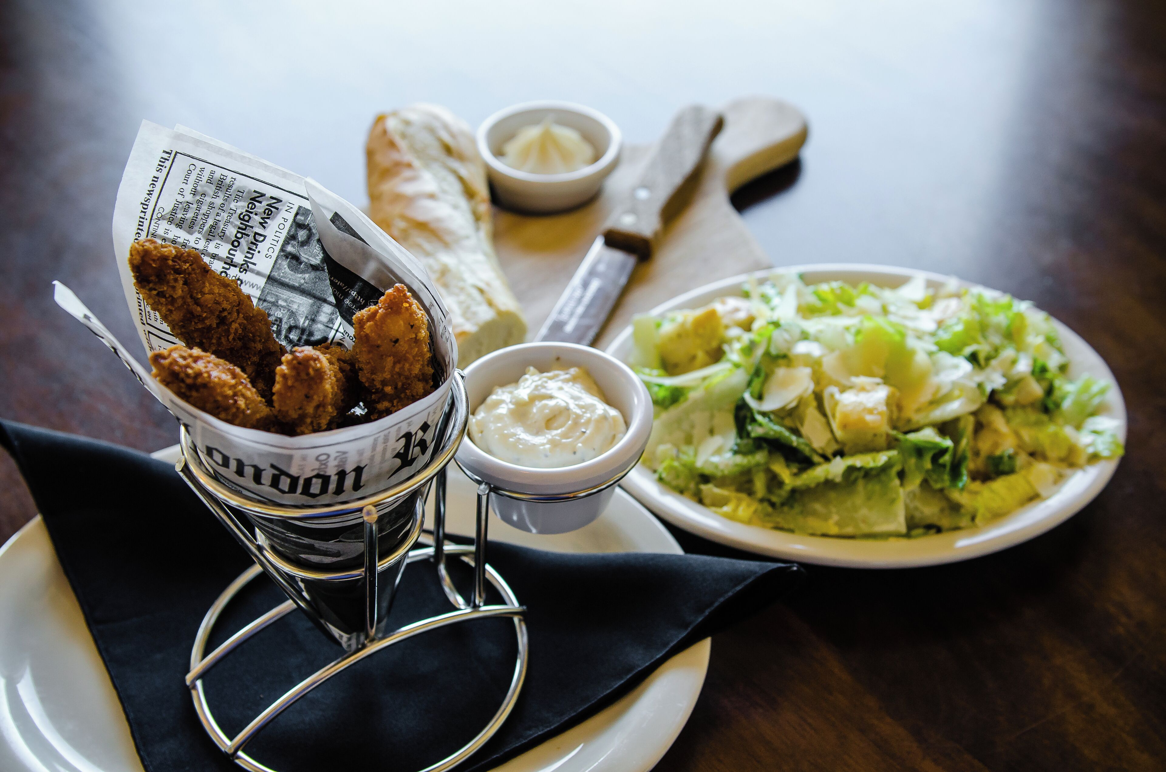 A plate with fried chicken, a small bowl of sauce, a piece of bread, and a salad on a wooden table.