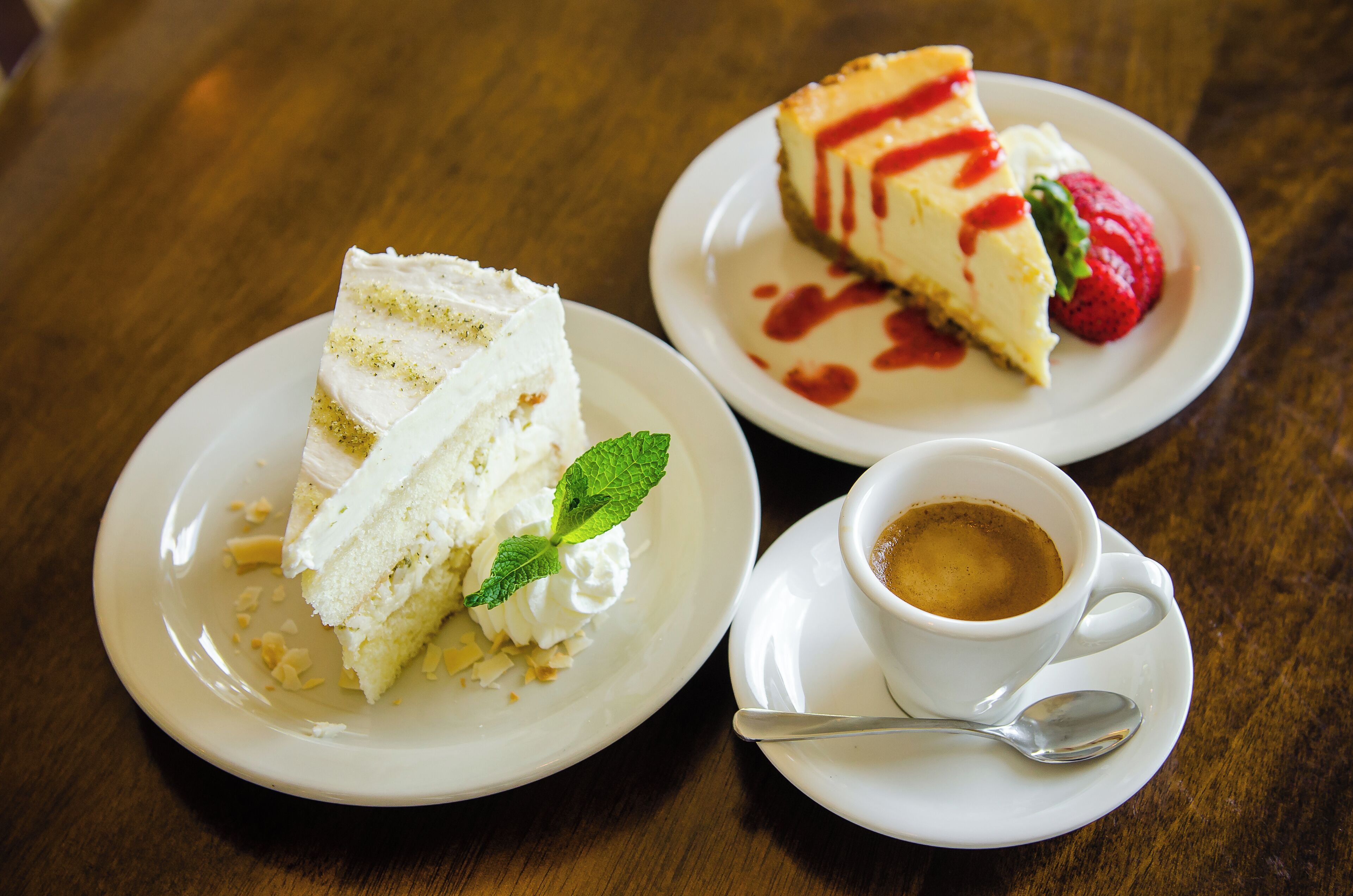Two white plates with slices of cheesecake and a cup of coffee on a wooden table.