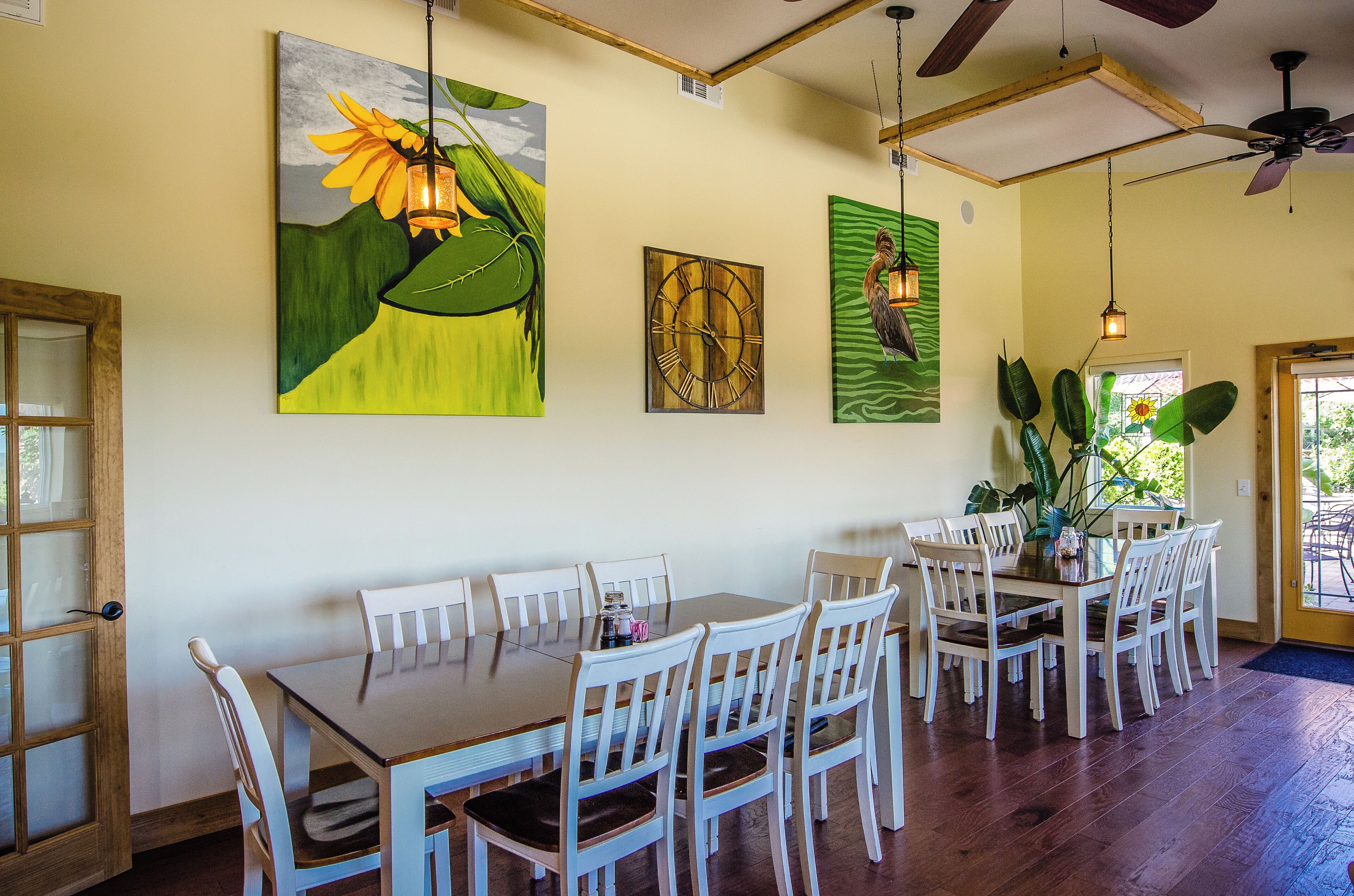 A dining area with a wooden table and white chairs, surrounded by artwork, and a ceiling fan.