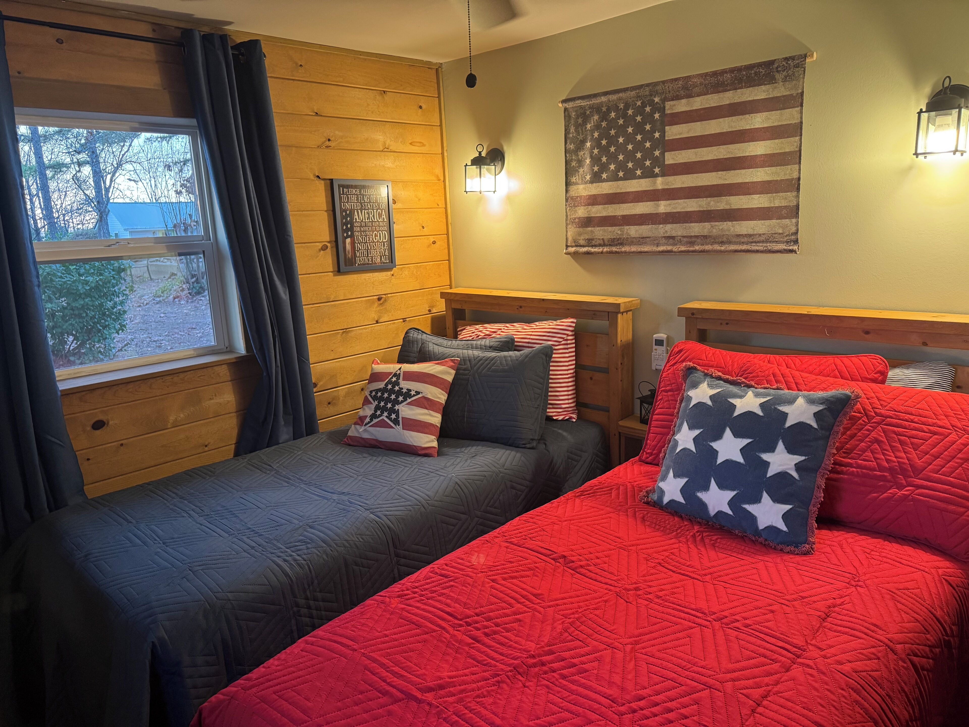 A room with two beds, one with a red quilt and the other with a dark quilt. There is an American flag on the wall, a window with curtains, and a wooden headboard.