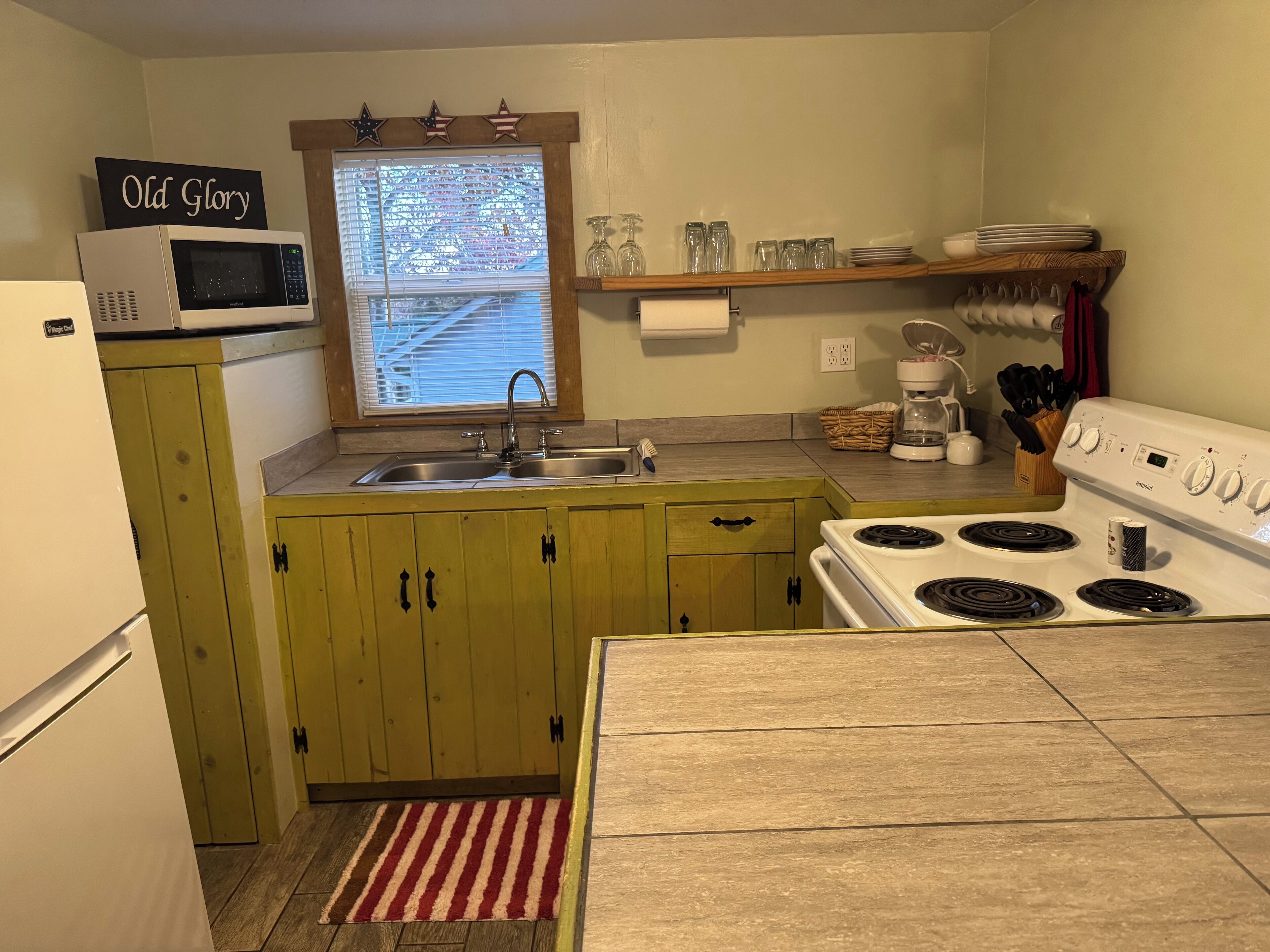 Una cocina compacta con microondas, lavamanos y estufa. Hay una ventana con persianas, un estante con cristalería y un cartel que dice "Old Glory".