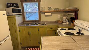 A compact kitchen with a microwave, sink, and stove. There is a window with blinds, a shelf with glassware, and a sign that reads "Old Glory".