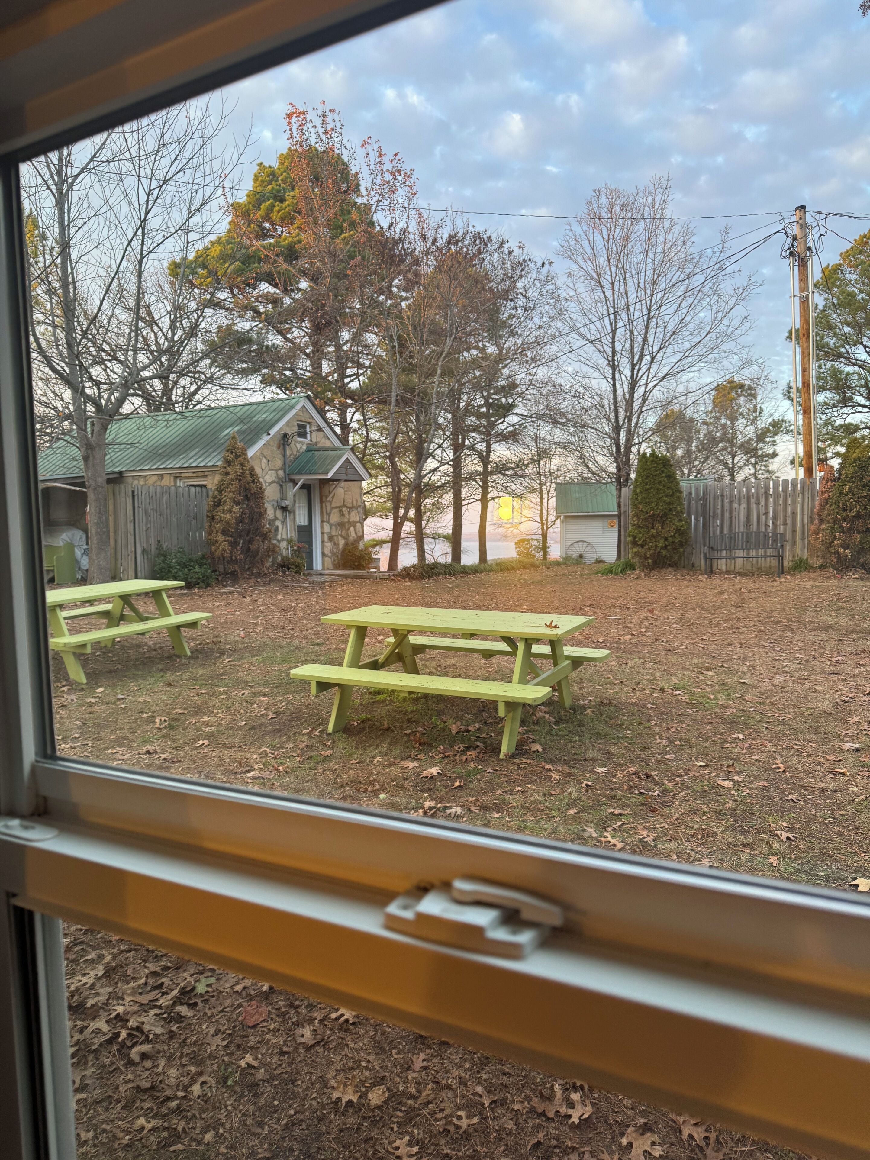 A view through a window of a backyard with picnic tables, a stone building, and a wooden fence.