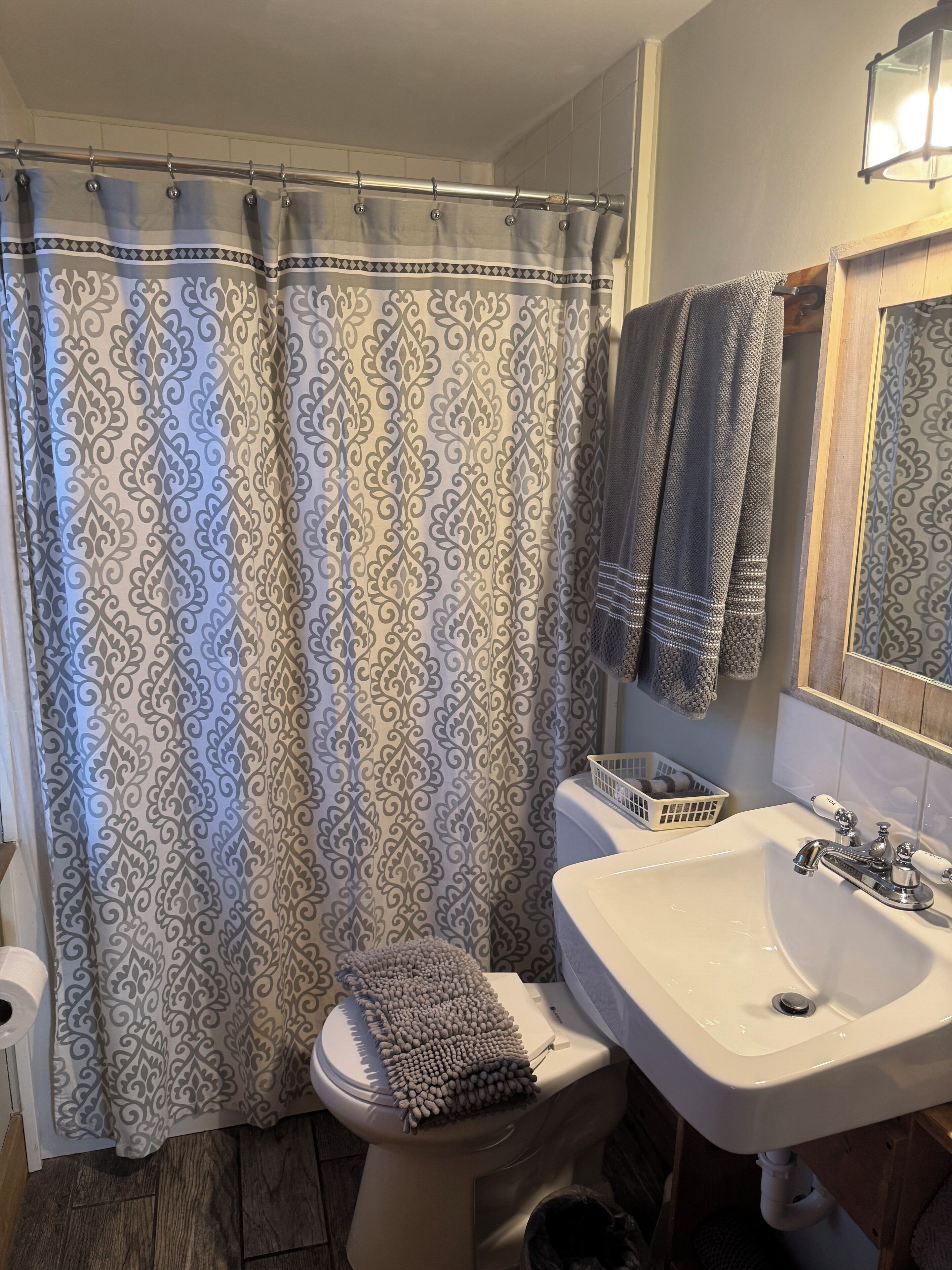 Une salle de bain avec un rideau de douche à motifs, des toilettes avec une serviette à portée de main, un lavabo avec un miroir et un panier fixé au mur.