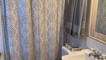 Une salle de bain avec un rideau de douche à motifs, des toilettes avec une serviette à portée de main, un lavabo avec un miroir et un panier fixé au mur.