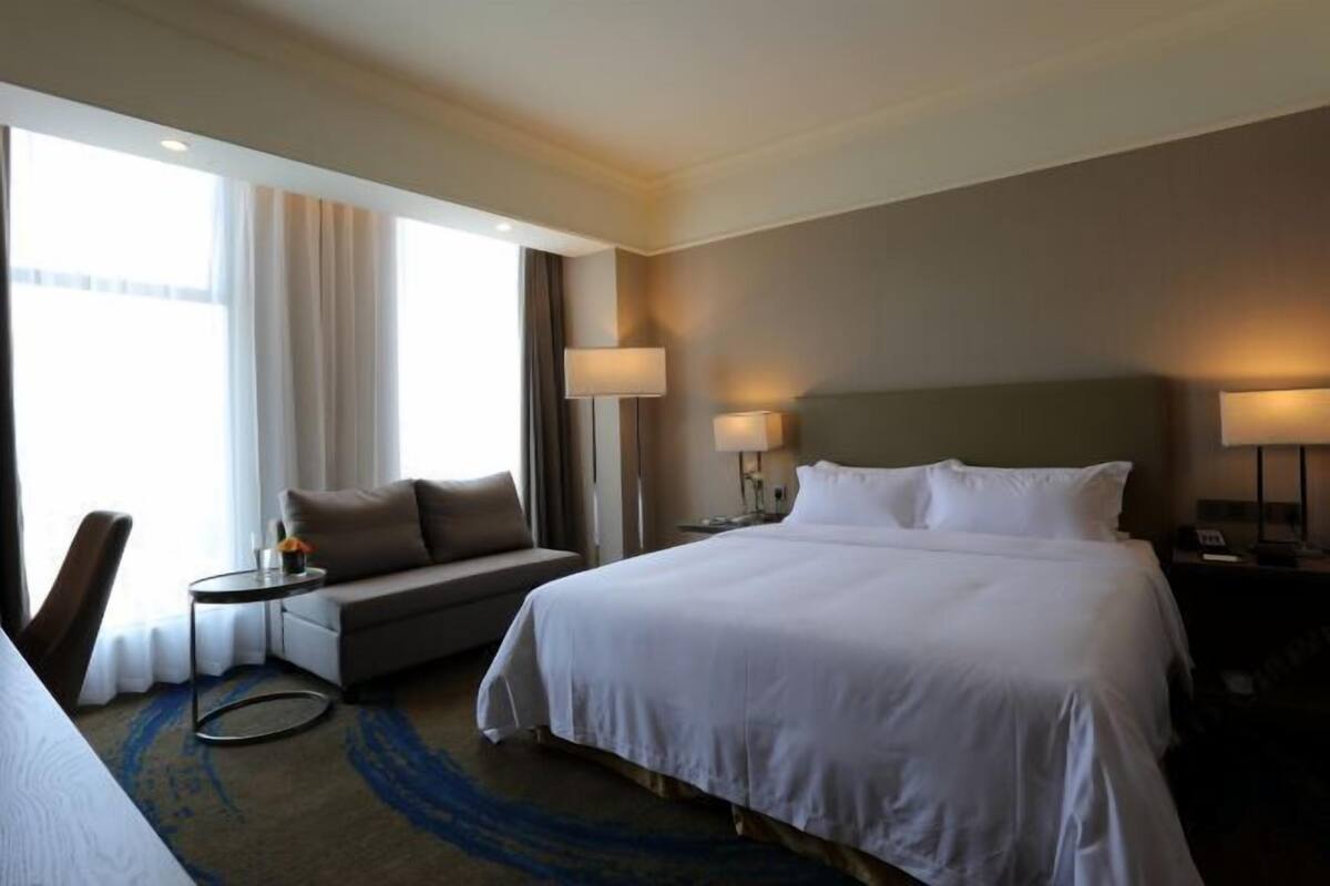 Una habitación de hotel con una cama grande, un sofá, una mesita y una silla. Hay dos lámparas en las mesitas de noche y una ventana con cortinas.