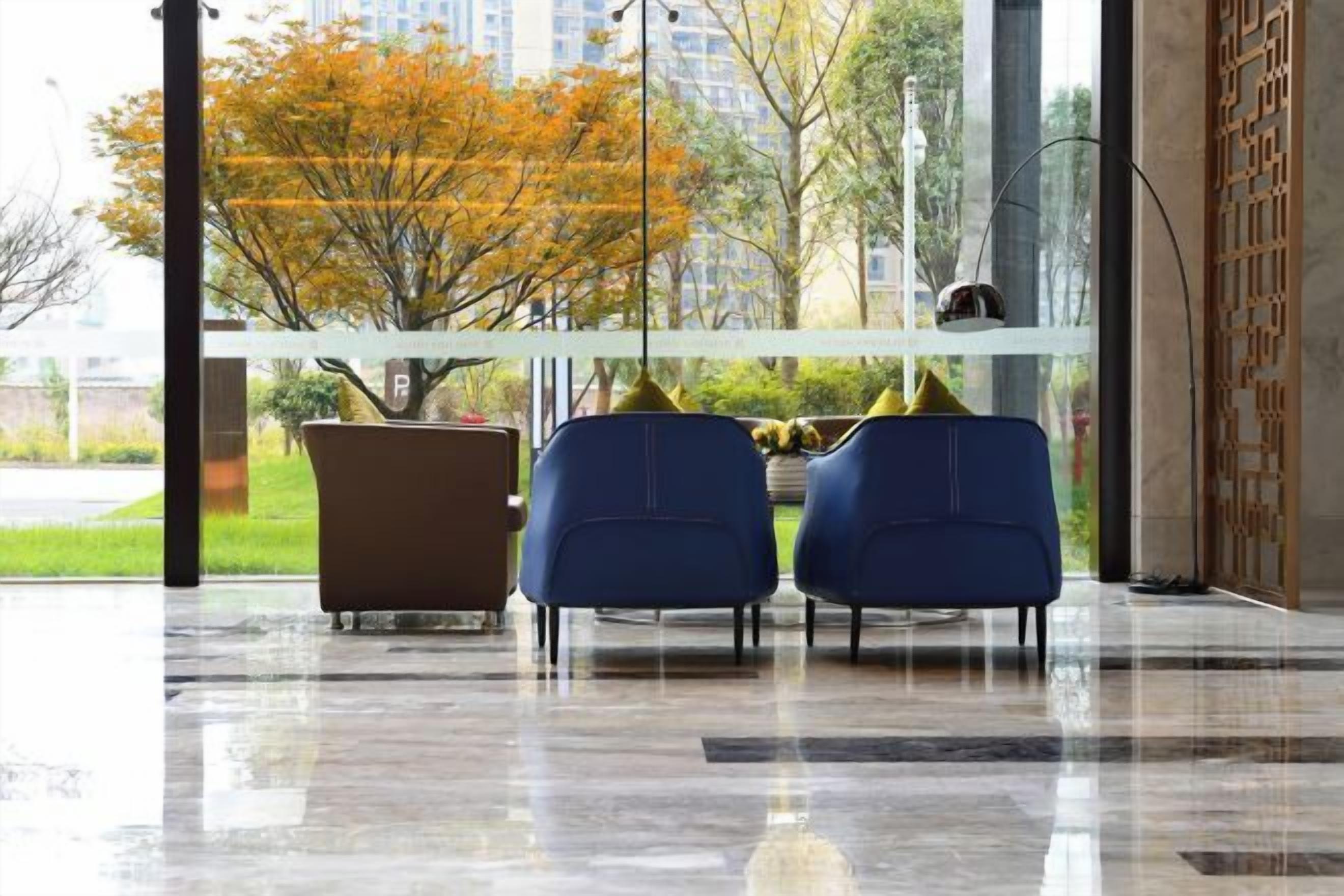 A waiting area with a view of trees and buildings, two blue chairs, a brown chair, and a glass partition.