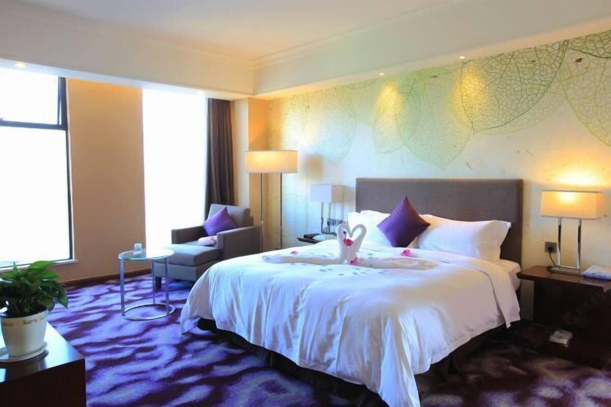 Habitación de hotel con una cama grande, dos mesitas de noche, una silla, una mesita pequeña y una planta en maceta.