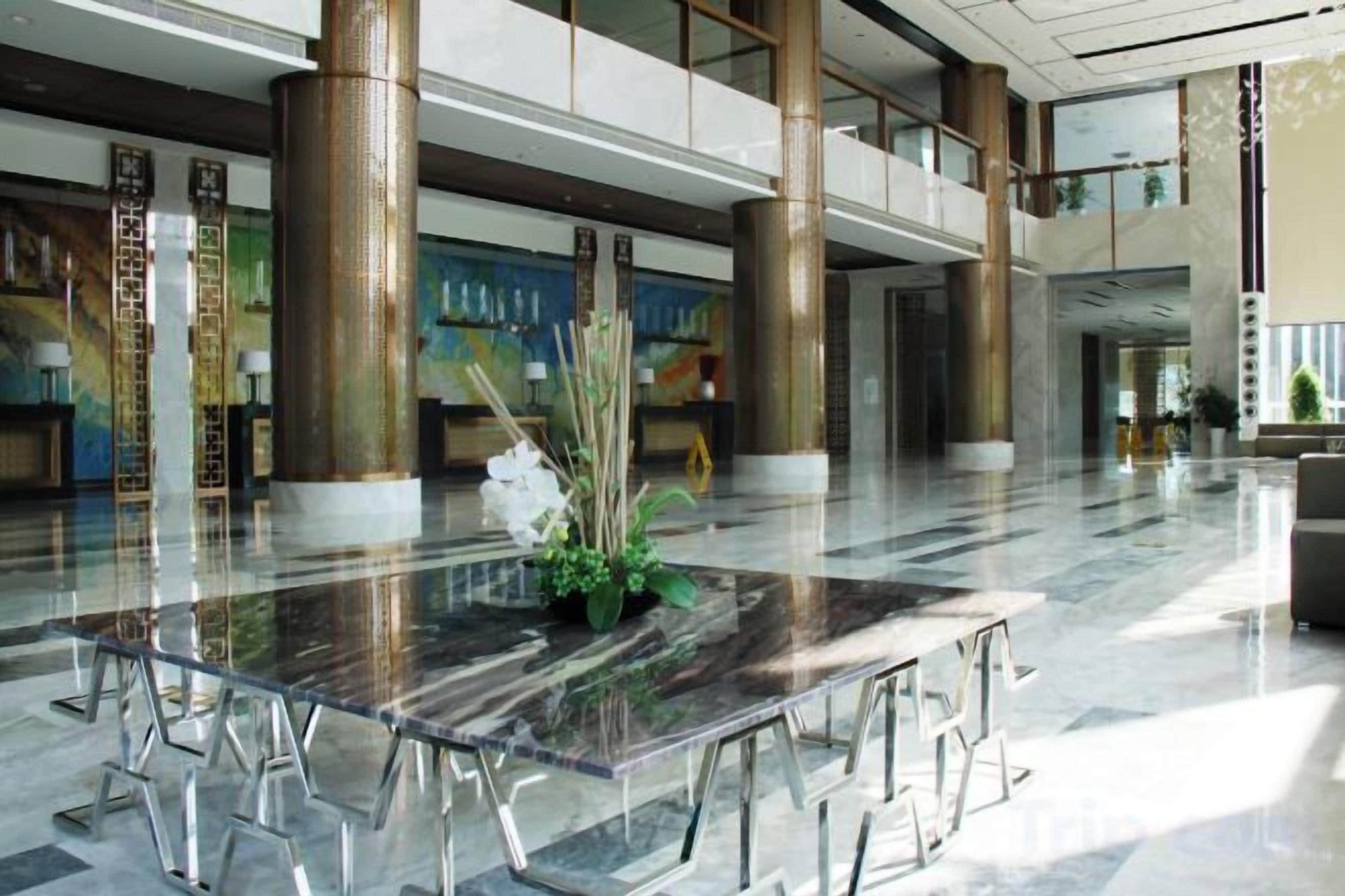 Un amplio vestíbulo con una mesa de vidrio en el centro, techos altos y grandes columnas.