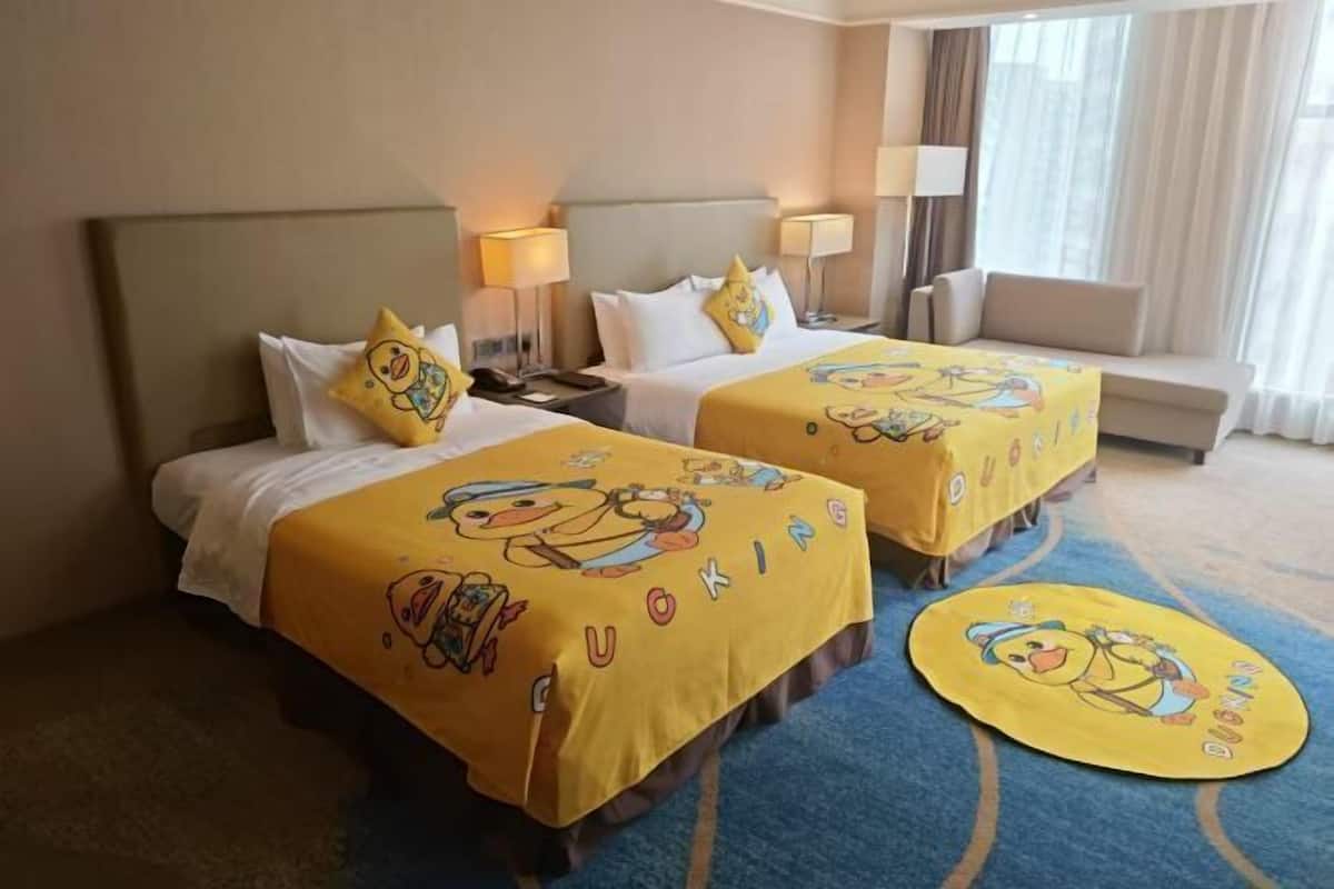 Habitación de hotel con dos camas, cada una con cubrecama y almohadas con motivos de patos amarillos. El suelo es de moqueta azul y tiene una alfombra redonda con diseño de patos.