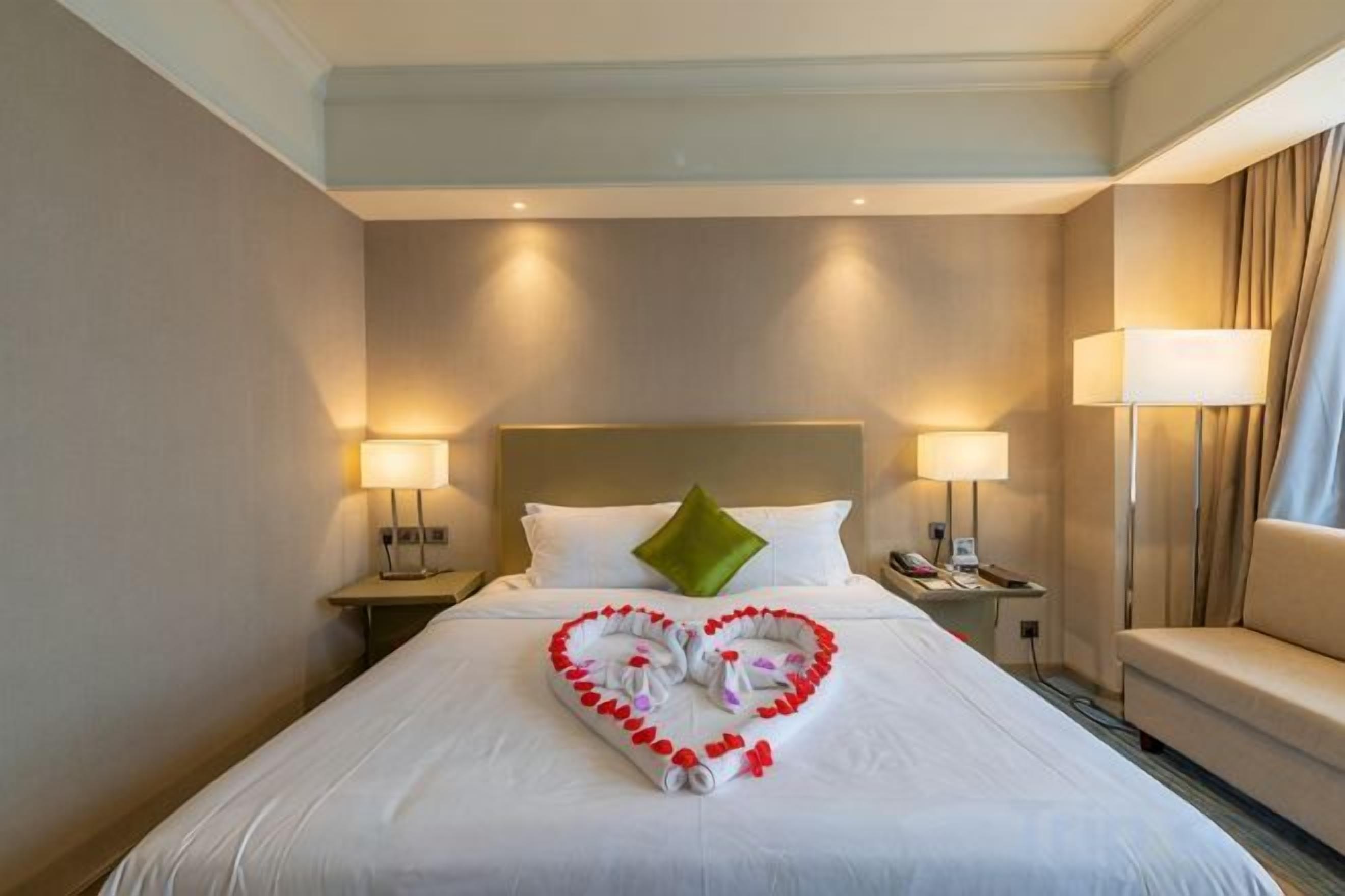 A hotel room with a neatly made bed, two bedside lamps, a green pillow, and a heart-shaped decoration on the bed.