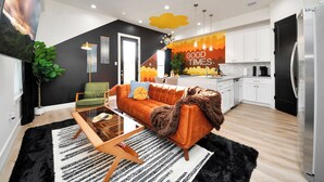A modern living room with a sofa, coffee table, and a TV. There is a kitchen area with white cabinets and a refrigerator.