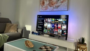 Olohuoneessa on litteänäyttöinen televisio, jossa näkyy elokuvavalikoita, harmaa sohva, koriste-esineillä varustettu sohvapöytä ja seinälle asennettu ilmastointilaite.