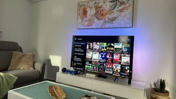 Een woonkamer met een flatscreen-tv waarop een menukaart met films te zien is, een grijze bank, een salontafel met decoratieve objecten, en een wandgemonteerde airconditioner.