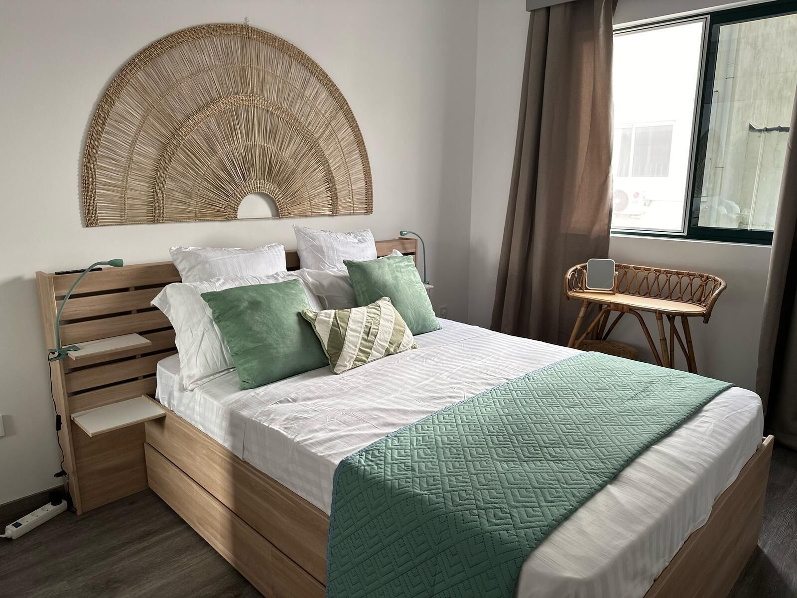 Un lit bien fait, avec une couette verte, une tête de lit en bois et une table de chevet surmontée d’une lampe.