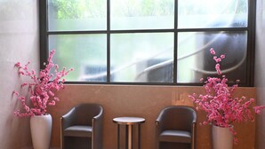 Zona salotto con due poltrone e un tavolino, affiancata da due grandi vasi con piante a fiori rosa, di fronte a un'ampia finestra.