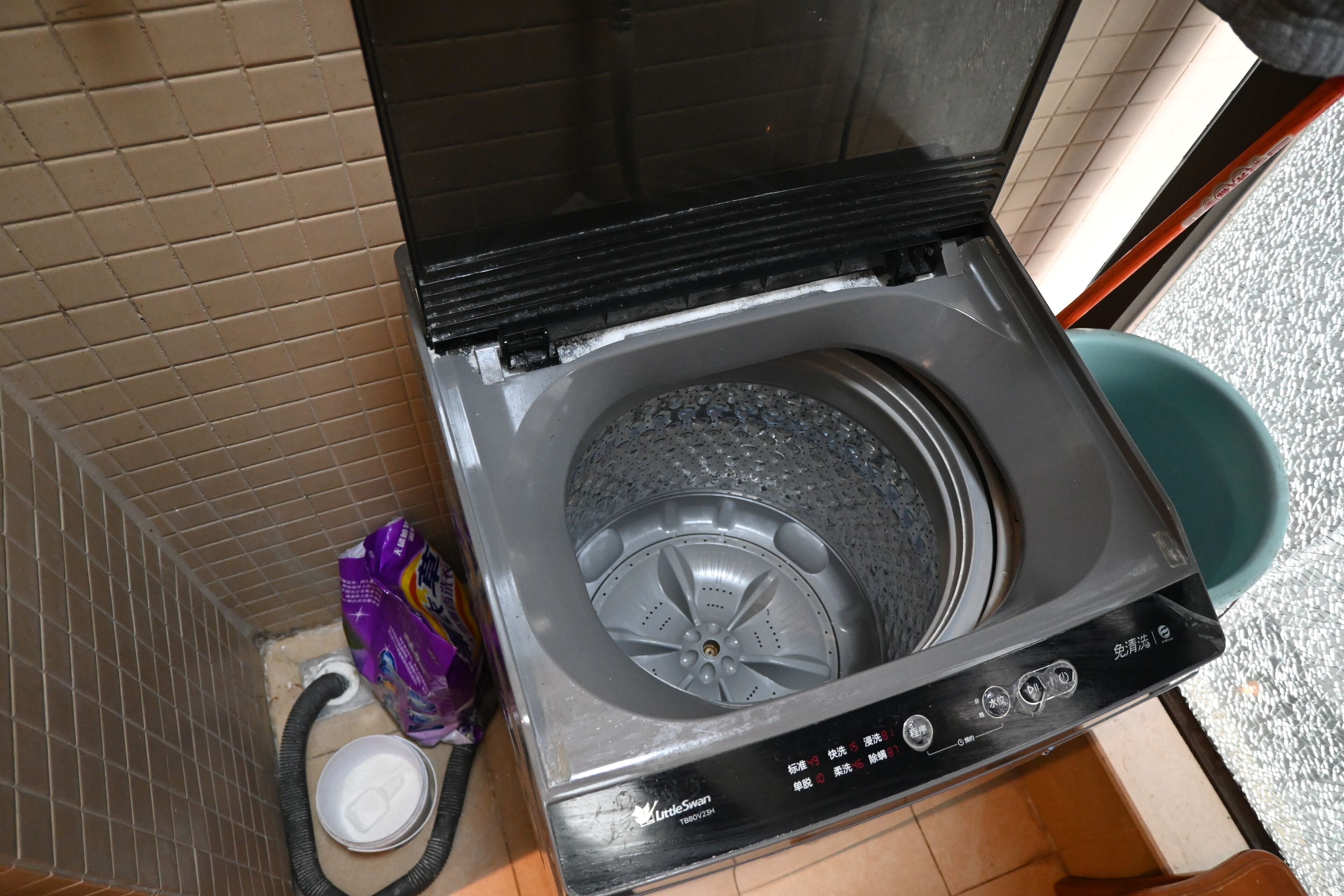 A compact, modern washing machine with a control panel, situated in a small laundry room with tiled walls and a visible water inlet hose.