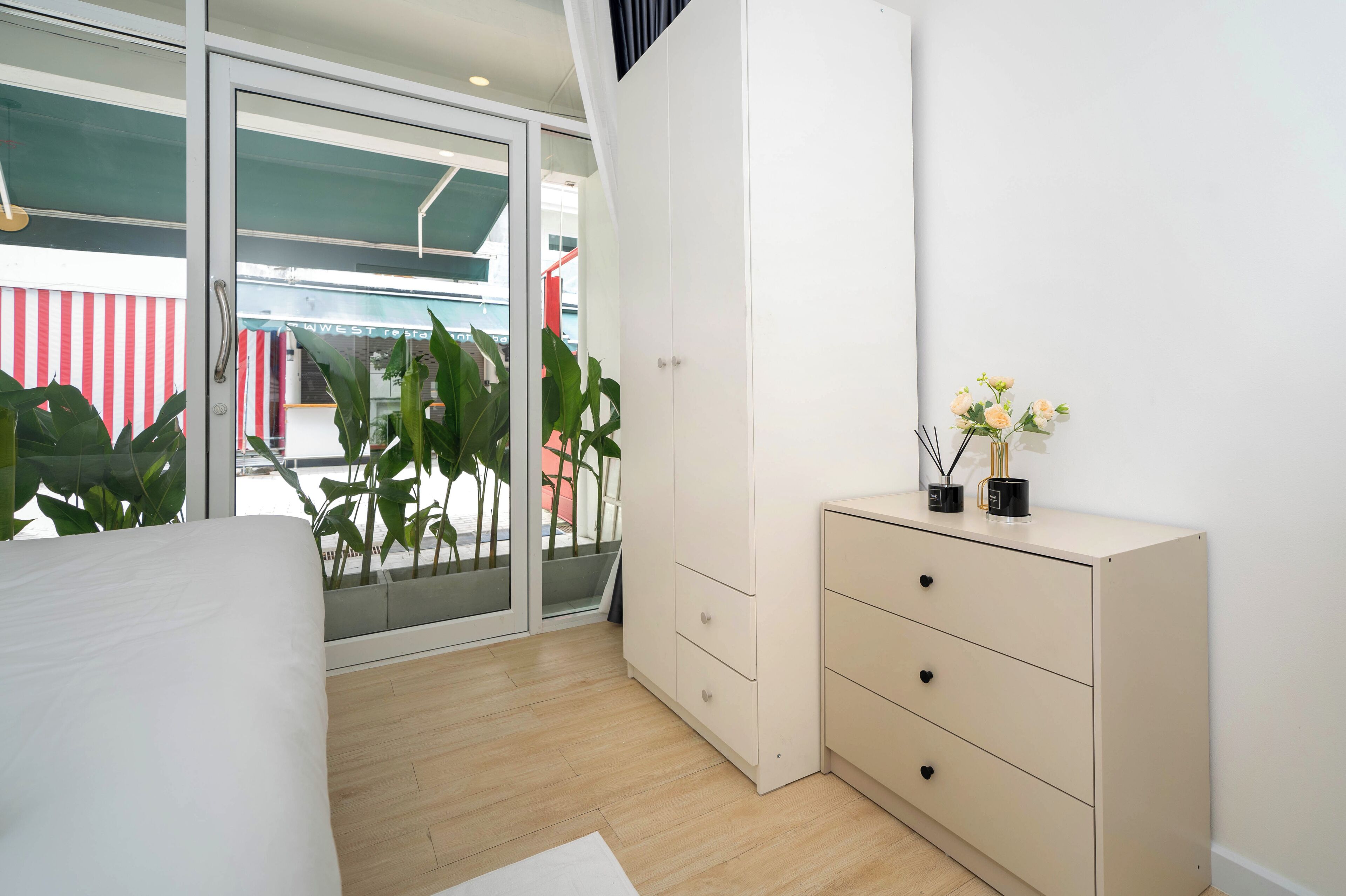 A modern bedroom with a bed, a sliding glass door leading to a balcony, a white dresser, and a potted plant.